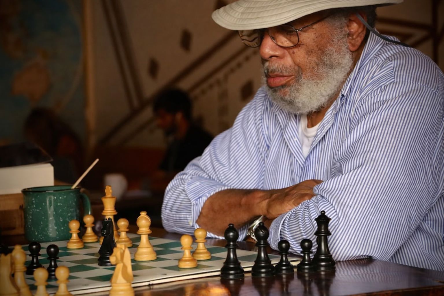 Äldre man med hatt och skägg spelar schack vid ett bord.