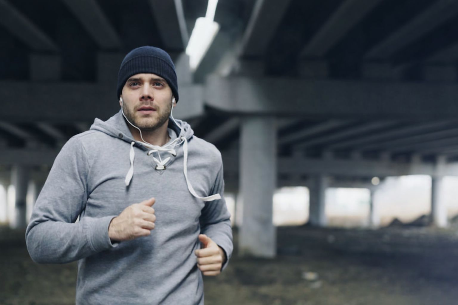 Man i mössa och grå hoodie joggar under en bro med hörlurar.